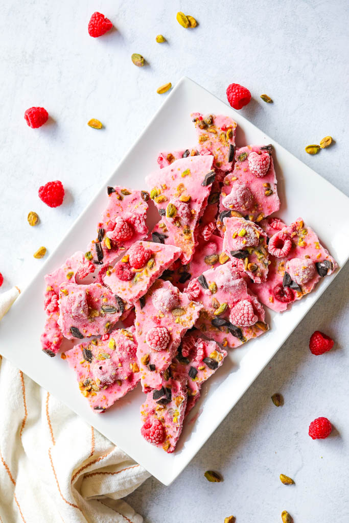 Pink raspberry frozen yogurt bark on a white plate with raspberries, cacao nibs and pistachios