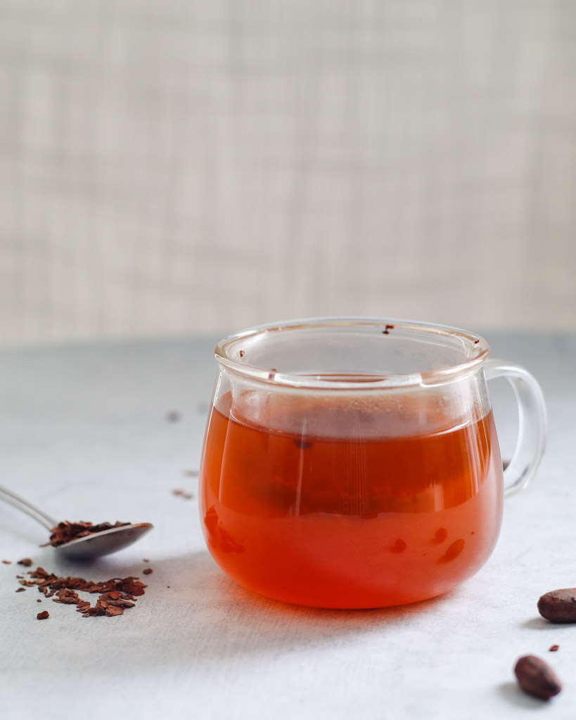 a clear mug filled with cacao tea with a spoon and scattered cacao nibs surrounding it
