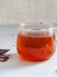 a clear mug filled with cacao tea with a spoon and scattered cacao nibs surrounding it