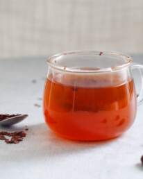 a clear mug filled with cacao tea with a spoon and scattered cacao nibs surrounding it