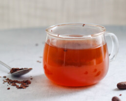 a clear mug filled with cacao tea with a spoon and scattered cacao nibs surrounding it