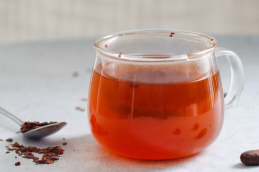 a clear mug filled with cacao tea with a spoon and scattered cacao nibs surrounding it