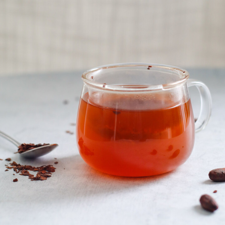 a clear mug filled with cacao tea with a spoon and scattered cacao nibs surrounding it